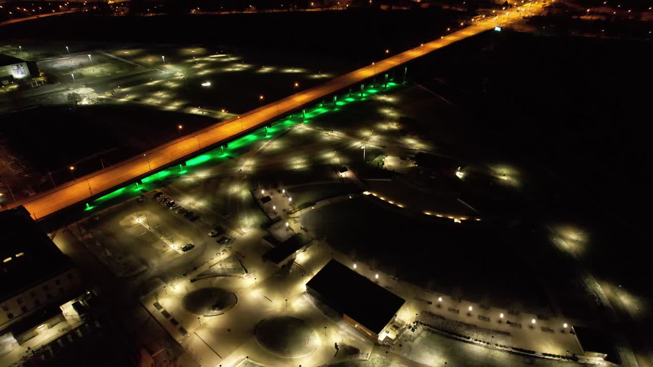 High-angle drone view of a glowing green-lit bridge over a city lot in Youngstown, Ohio. Urban geometry and parking lights stand out in this cinematic night scene.