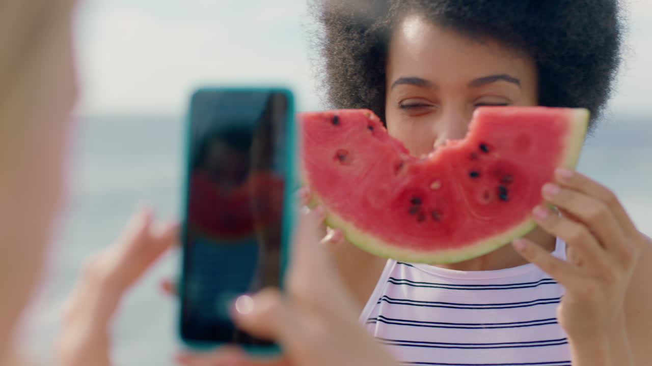 hermosa mujer con afro comiendo sandía en la playa posando para una amiga tomando fotos usando un teléfono inteligente amigas compartiendo el día de verano en las redes sociales divirtiéndose en la playa 4k