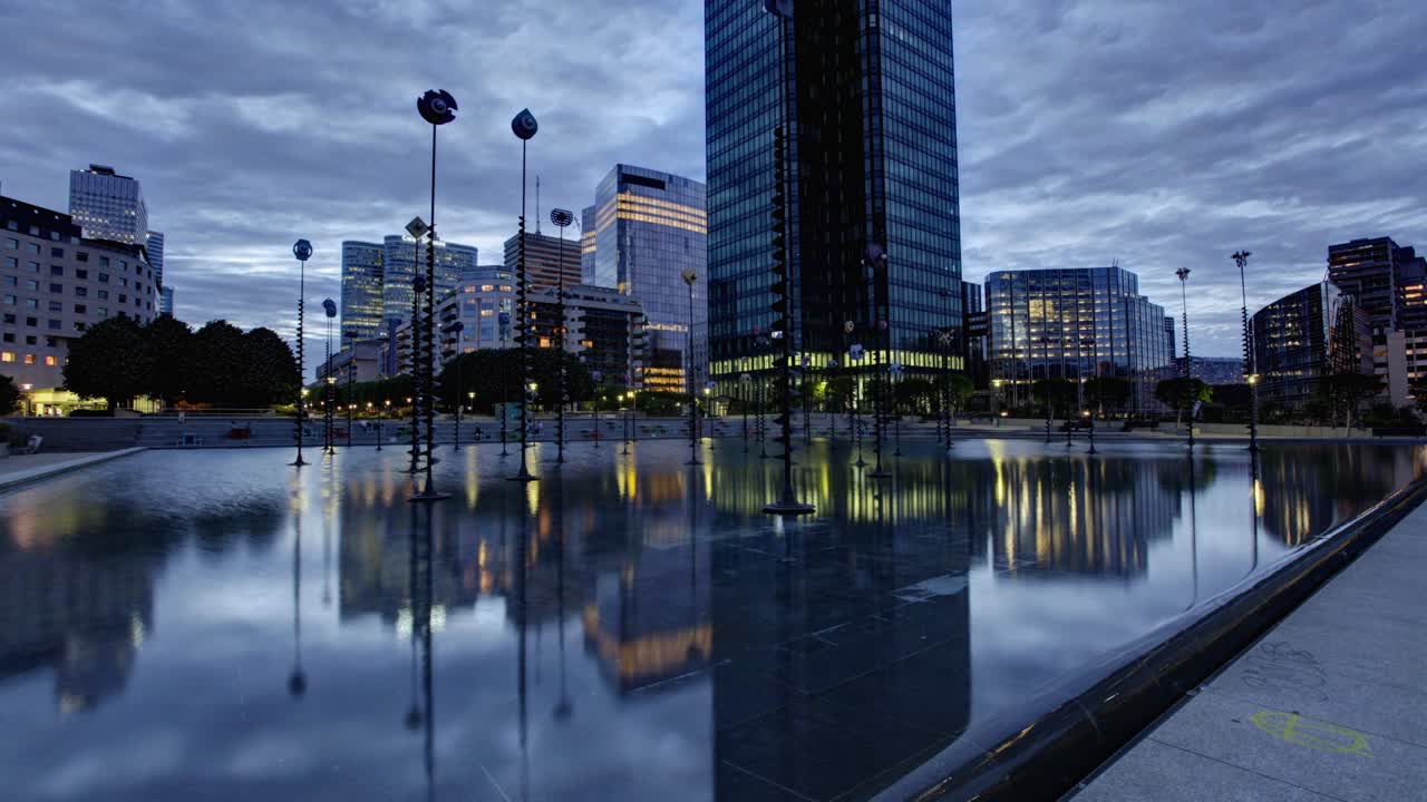 Timelapse showcases a modern cityscape at dawn, boasting tall glass and steel buildings reflected in a serene water feature, encircled by globe street lamps under a clear pale sky