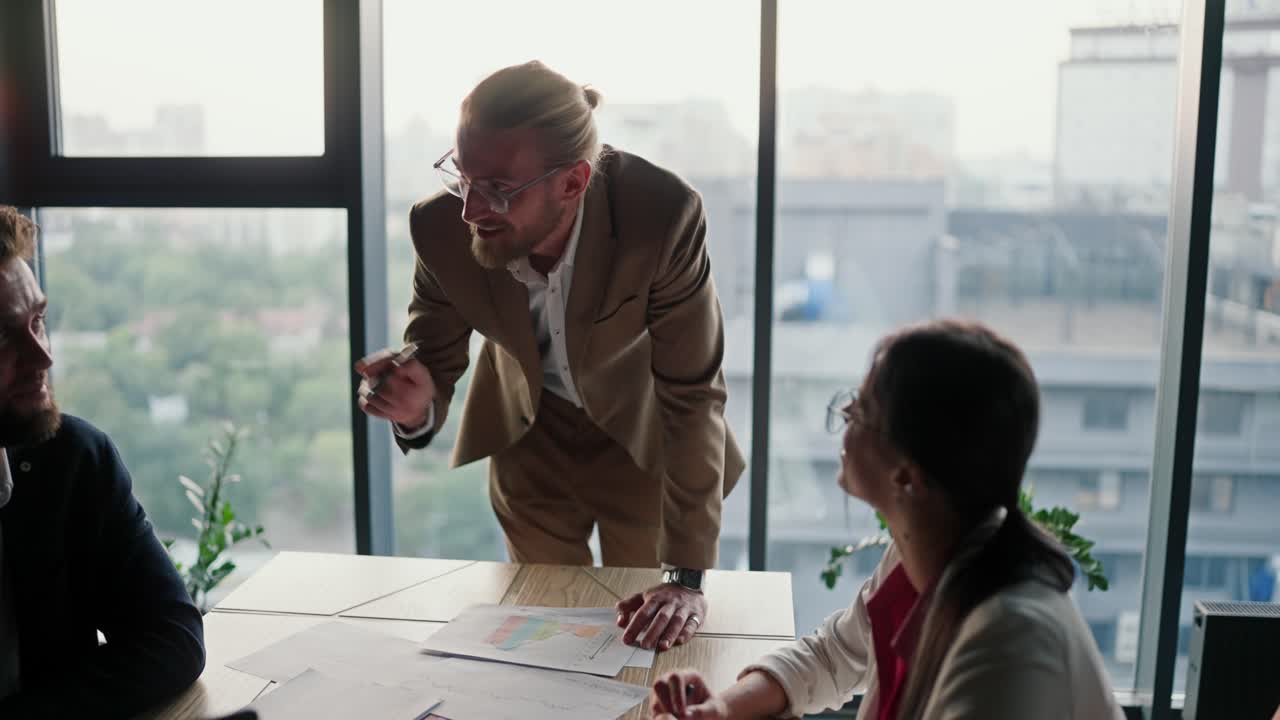 Zoom in: Young blond boss in glasses with a light brown beard with his employees leaning on the table in a modern office with a panoramic window