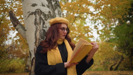 contemplative young lady goes through her book, seated alone in park, about to open another page, wearing yellow beret and muffler, background features trees with golden autumn leaves