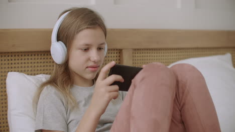 school girl is viewing funny video on display of smartphone and listening by headphones in her bedroom watching cartoons and films