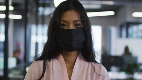 portrait of mixed race businesswoman wearing face mask standing in office looking to camera