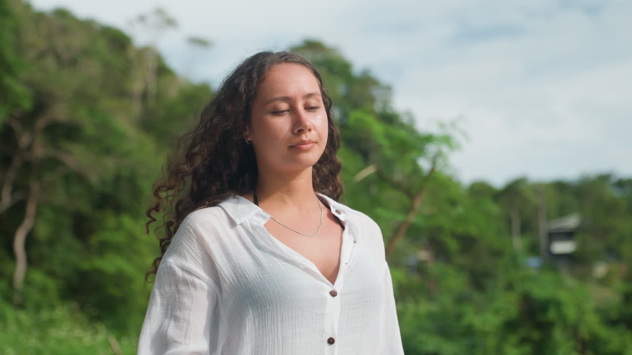 Donna su una verde collina dell'isola, che indossa una camicetta bianca, capelli ricci, che gira lentamente la testa e chiude gli occhi per inalare la calda brezza tropicale. Ritratto spontaneo che cattura un'atmosfera contemplativa, fogliame lussureggiante, distante.