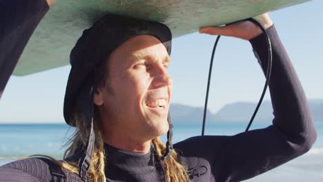 video de un hombre caucásico sonriente con rastas llevando una tabla de surf en la cabeza en una playa soleada