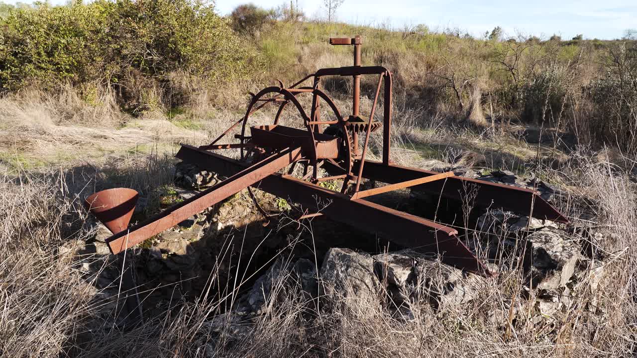 Primo piano di una vecchia pompa meccanica, macchiata di ruggine e tempo, accanto al pozzo di una fattoria; la sua struttura sbiadita riecheggia anni di utilizzo, un tempo fondamentale per attingere acqua per sostenere i raccolti e la vita rurale.