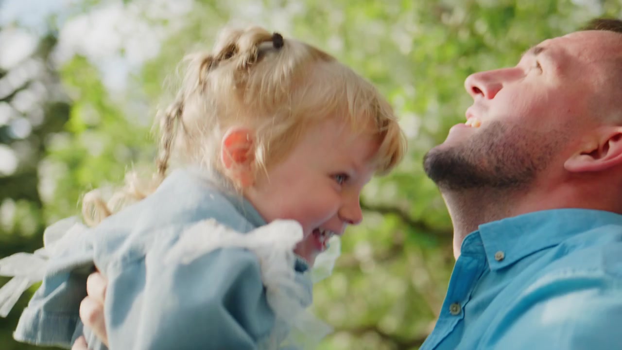 Uomo tiene in braccio un bambino che ride all'aperto. Il bambino guarda in alto con gioia. L'uomo ricambia il sorriso calorosamente. Alberi verdi e cielo sullo sfondo. Perfetto per temi familiari, genitorialità o natura. Tono luminoso e allegro