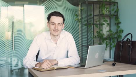 portrait of a handsome, young, stylish man in business attire sitting at a desk with a laptop in a modern office, talking and gesturing with his hands while looking into the camera