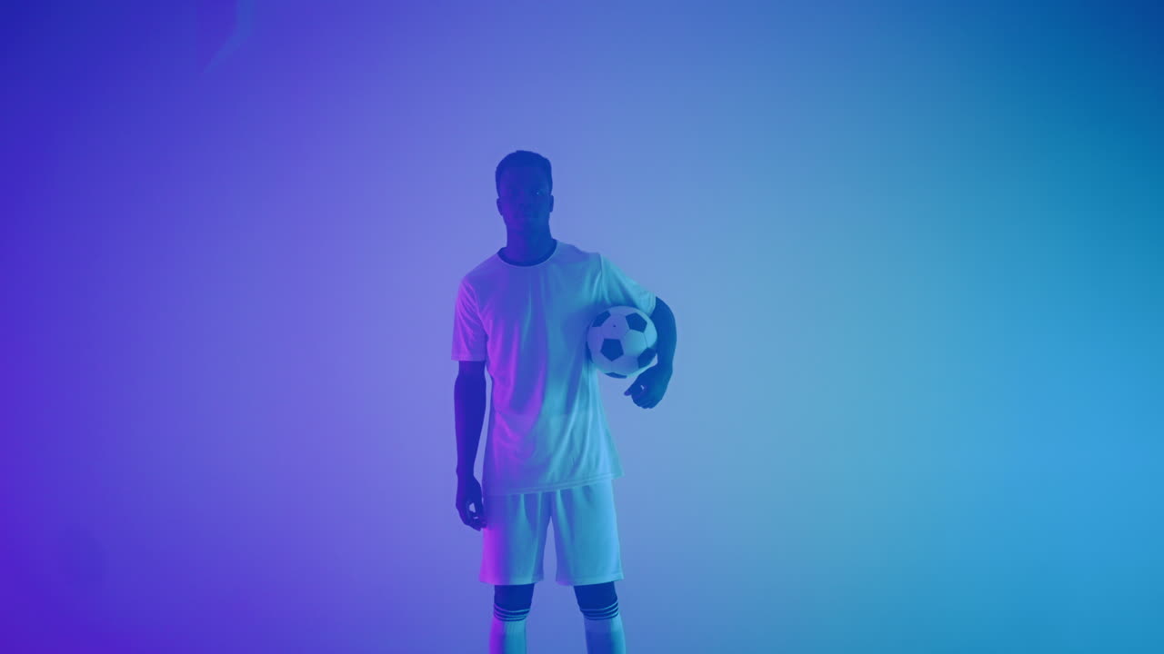 Ritratto al rallentatore di un giocatore di football americano professionista di colore in uniforme bianca, che guarda in camera sotto la luce neon blu-rossa dello studio. Un giocatore di football americano brutale, sicuro di sé e vittorioso