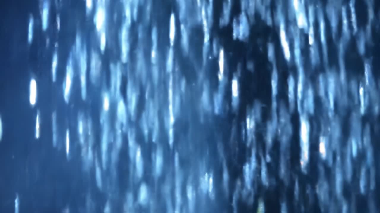 Raindrops fall continuously, creating a shimmering effect as they catch the light. The surroundings are dim, enhancing the delicate beauty of the falling water in the evening hours.