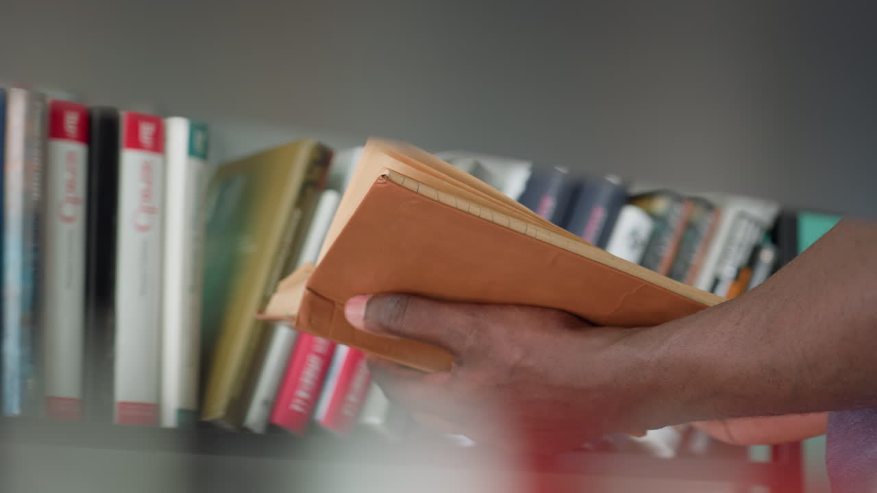 Blurred view of adult holding vintage brown notebook in hand near bookshelf filled with colorful books, creating focused foreground with soft background