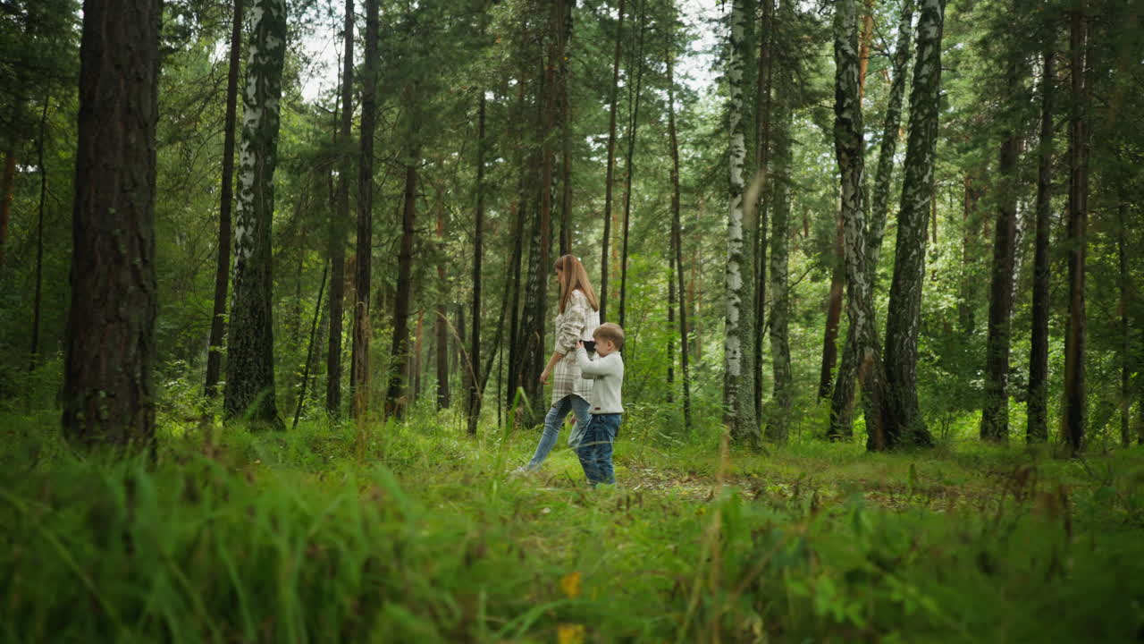 Side view of mother strolling through green forest with two children, enjoying nature walk as boy curiously holds microscope, surrounded by lush trees and tall grass