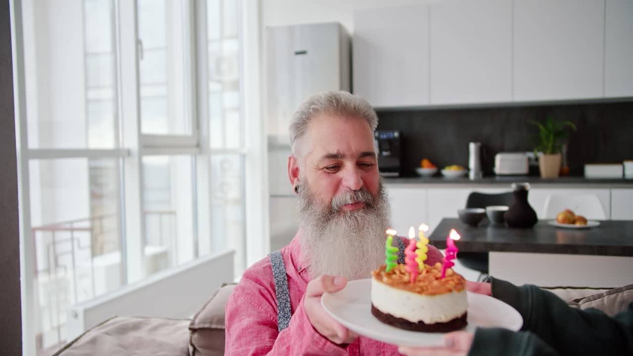 una chica morena con una chaqueta verde trae un pastel junto con velas para su padre anciano con cabello gris y una barba exuberante para su cumpleaños en un apartamento moderno