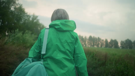 back view of an elderly woman in a green raincoat carrying a mint-colored bag, walking along a path surrounded by greenery and trees, under a soft, cloudy sky as she moves forward