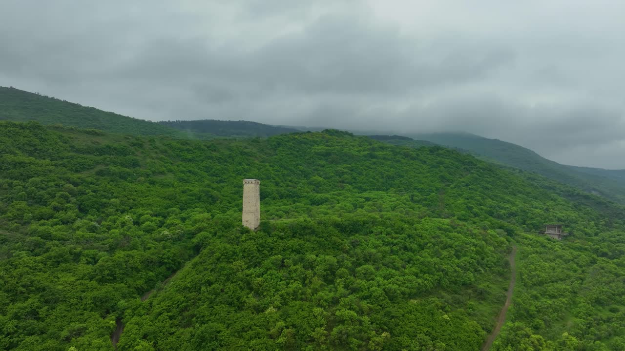 A solitary ancient stone tower stands proudly on a lush green ridge overlooking the misty hills of Tbilisi under a cloudy sky