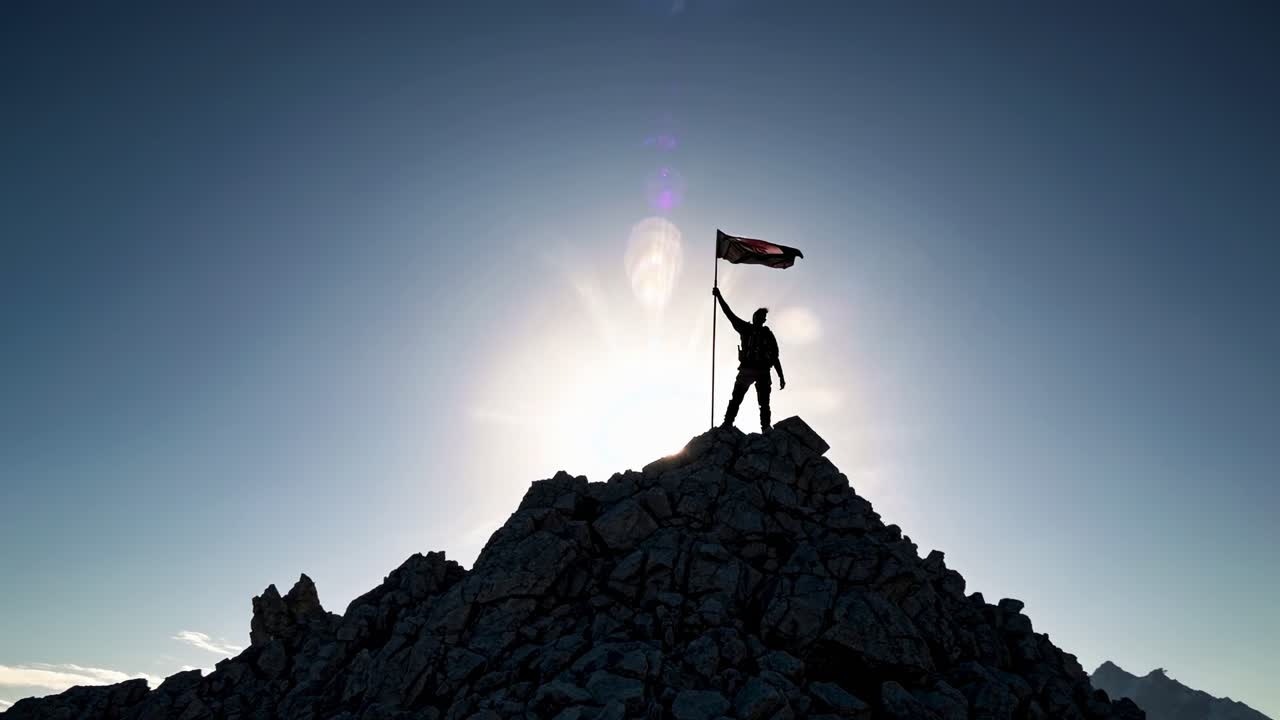 Silhouette of a person with a flag atop a rocky peak, captured from a low-angle, backlit by the sun