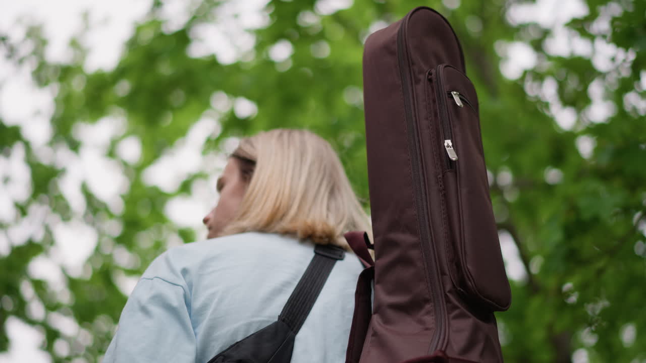 chitarrista caucasico biondo che trasporta una custodia per chitarra sulla spalla in un parco alberato, giacca di jeans, custodia imbottita marrone con dettagli a zip, prospettiva sopra la spalla, atmosfera tranquilla e riflessiva, preparazione