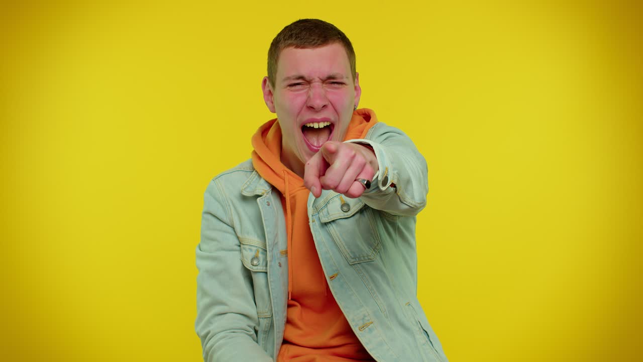 un joven con una chaqueta de vaqueros y una sudadera con capucha señala intensamente a la cámara con una expresión enojada.