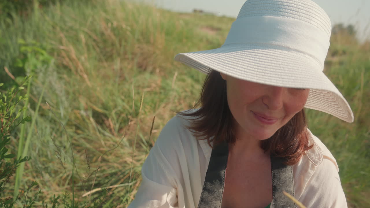 Overhead view of woman with camera squatting in tall grass wearing white sun hat, smiling softly while adjusting settings to capture wild plants in warm natural light during outdoor photography