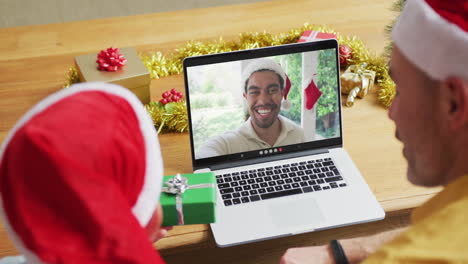 padre caucásico con hijo con sombreros de santa usando una computadora portátil para una videollamada de navidad, con el hombre en la pantalla