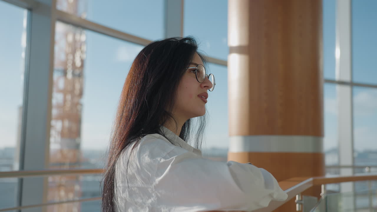 Calm woman with long dark hair and glasses rests on wooden rail in sunlit modern space, wearing white shirt and soft expression, looking ahead with warmth and gentle contemplation