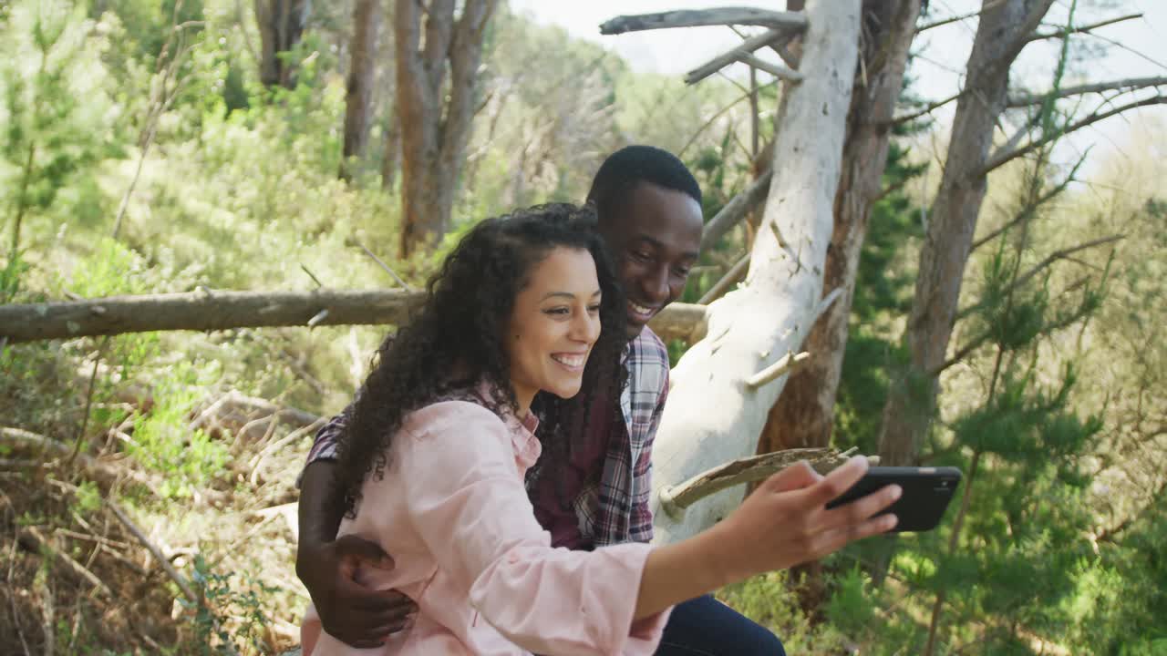 una pareja diversa sonriente tomando selfies y sentada en un árbol en el campo