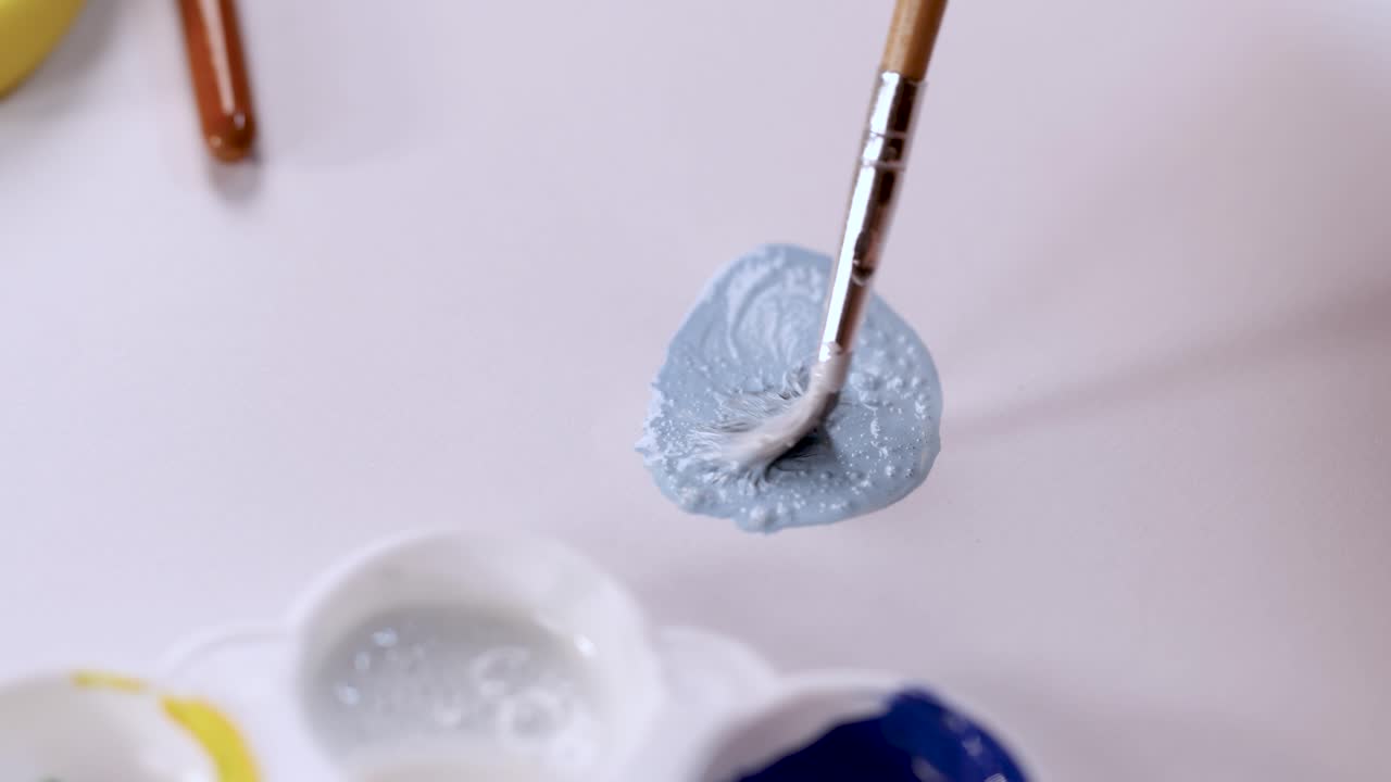 Paintbrush mixes light blue paint on palette in bright, soft lighting, overhead close-up view