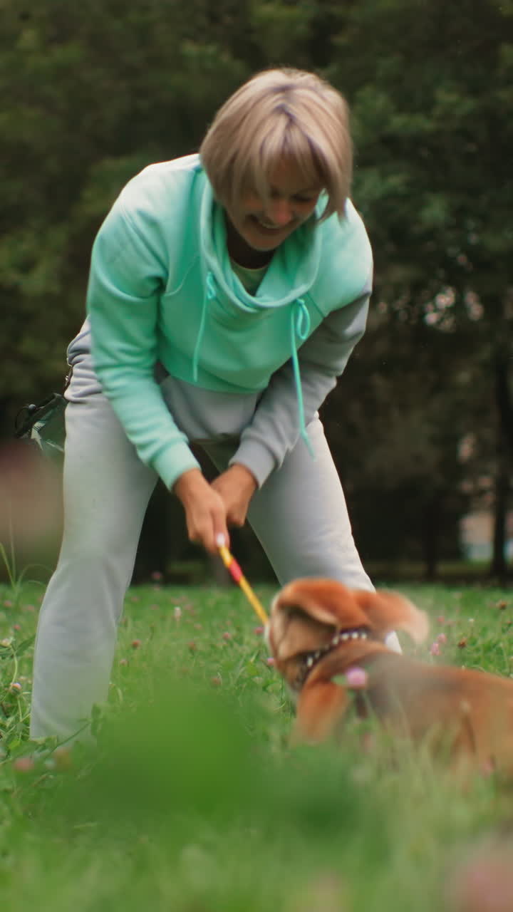 Mujer caucásica conectando con su perro al aire libre en un campo de tréboles, jugando y guiándolo con suavidad, figura de abuela que ofrece una interacción de estilo terapéutico, postura tranquila con correa y collar, árbol frondoso
