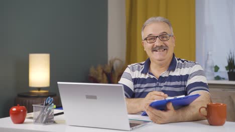 Home-office-worker-old-man-smiling-at-camera-looking-at-paperwork.