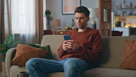 frowning man looking mobile phone screen on cozy couch. serious guy messaging