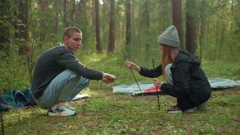 man squatting on forest floor assembling tent pole while lady kneels opposite handing over another pole during quiet camping moment surrounded by trees and scattered camping gear