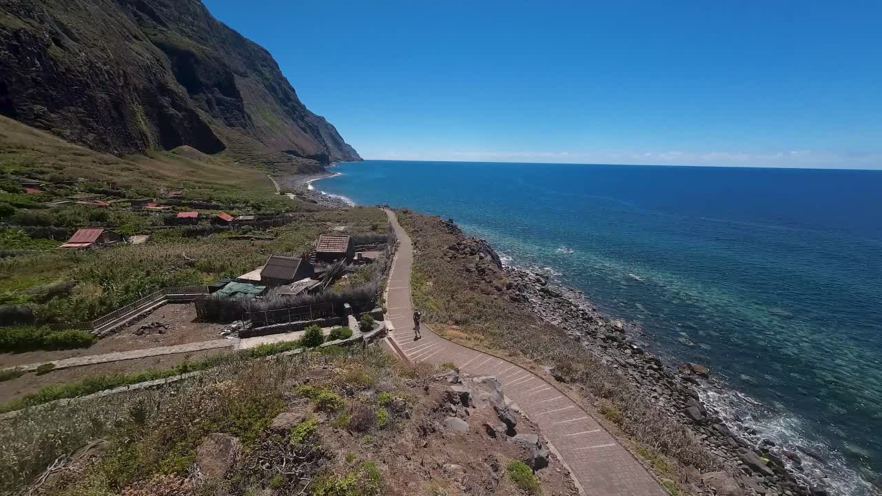 Small homes and farms dot the coastal landscape of Madeira Island. People enjoy a walk along the shoreline path with stunning blue sky and sea. Aerial view from drone.