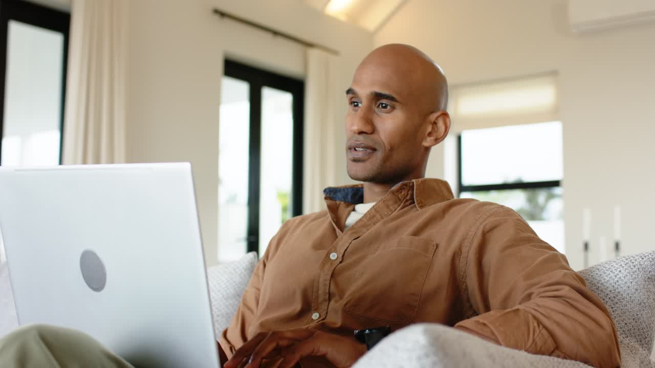 Man viewing laptop screen, checking time on smartwatch and typing to respond on sofa at home
