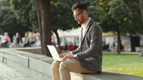 Apuesto-Hombre-De-Negocios-Sentado-En-La-Pared-En-El-Parque-De-La-Ciudad-Y-Trabajando-En-Su-Computadora-Portátil