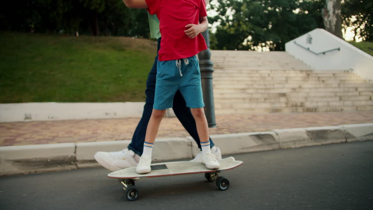Close-up shot of a father in blue jeans helping his little son in a red T-shirt ride a skateboard in the park. Little boy rides a white skateboard along the road in the park