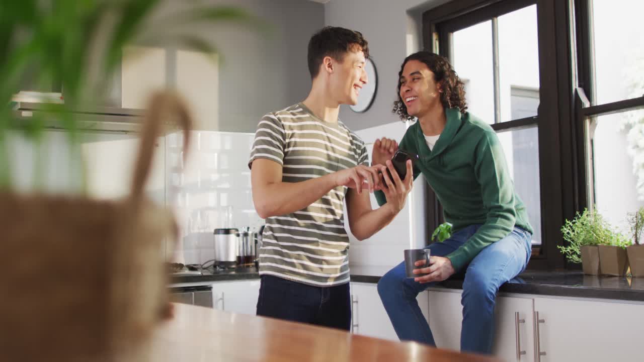 feliz pareja masculina diversa bebiendo café y usando un teléfono inteligente en la cocina