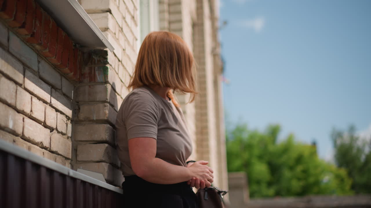 Office clerk with light brown hair lean against sunlit brick wall holding handbag, adjusting dark sunglasses while glancing right and left, sunlight casting shadows on face
