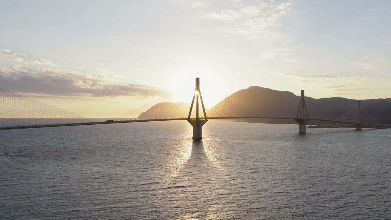 Panoramica drone del Ponte Rio-Antirrio sagomato al tramonto. Il sole scompare dietro i piloni per poi riemergere. Mare calmo, riflessi dorati.