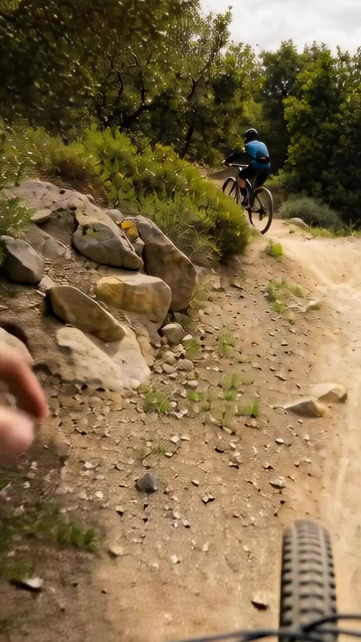 Vídeo vertical: Seguindo a roda da bicicleta e uma mão apontando enquanto um ciclista de camisa azul sobe uma curva na trilha.