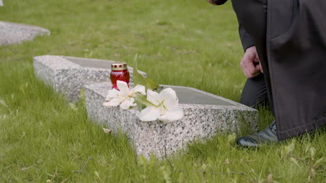 hombre irreconocible con traje negro arrodillado y poniendo flores y una vela en una lápida en un cementerio 1