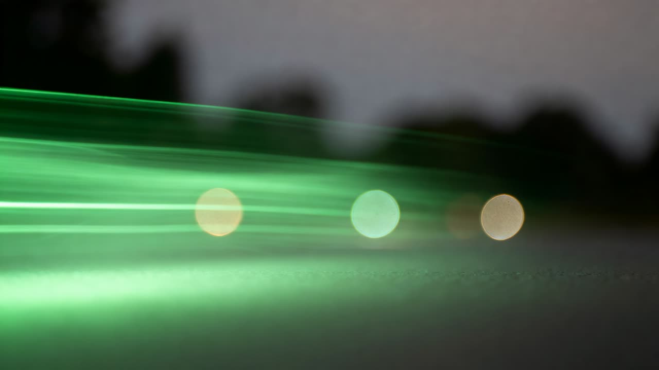 Moving green light sweeping past treeline, creating bands and passing three bokeh lights over road