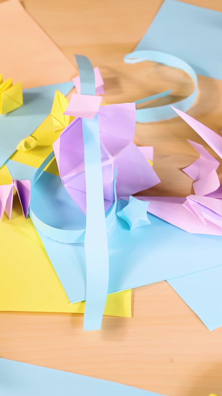Hands fold pastel origami paper into birds and boats on wooden table, bright overhead lighting