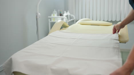 body work therapist in navy uniform sprays disinfectant onto cotton pad and wipes down cream massage chair in bright white treatment room, ensuring sterile, inviting setup for client relaxation