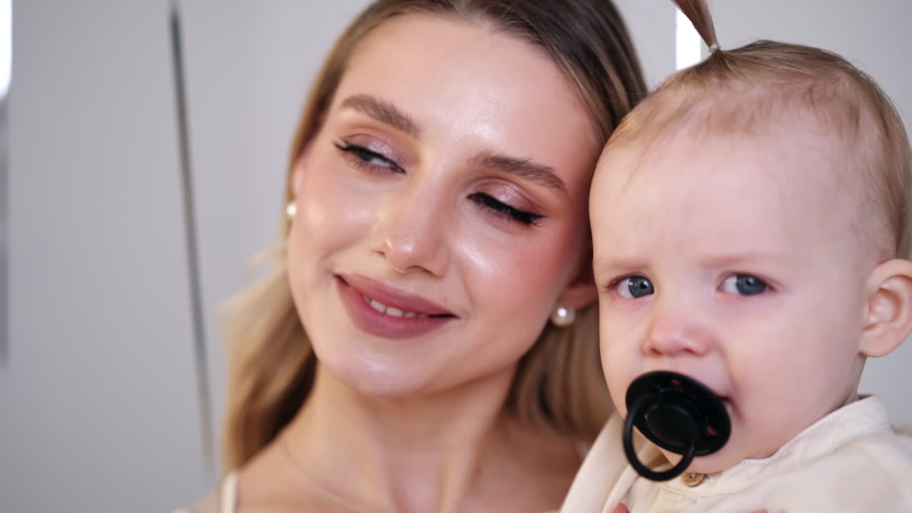 Beautiful Caucasian young mom holding her baby. Cute child with pacifier in mouth looks at balloons around. Close up.