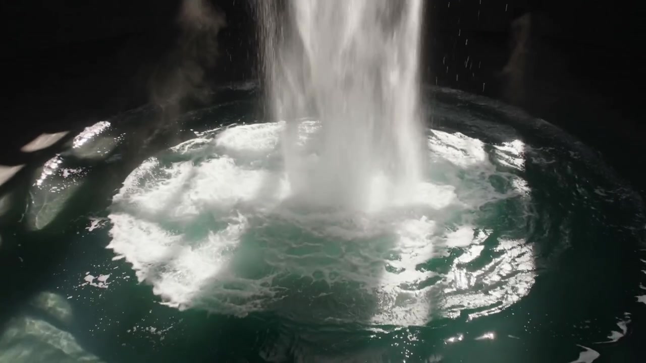 Colonna d'acqua che cade producendo un nucleo schiumoso e increspature in una vasca circolare, con riflessi verdi.