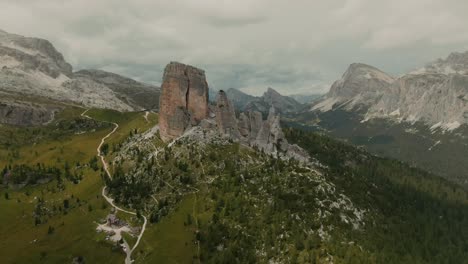 aerial view aproaching massive rock formations with distant tall mountains in the background, green fields at the bottom, cloudy day, cinematic color grade