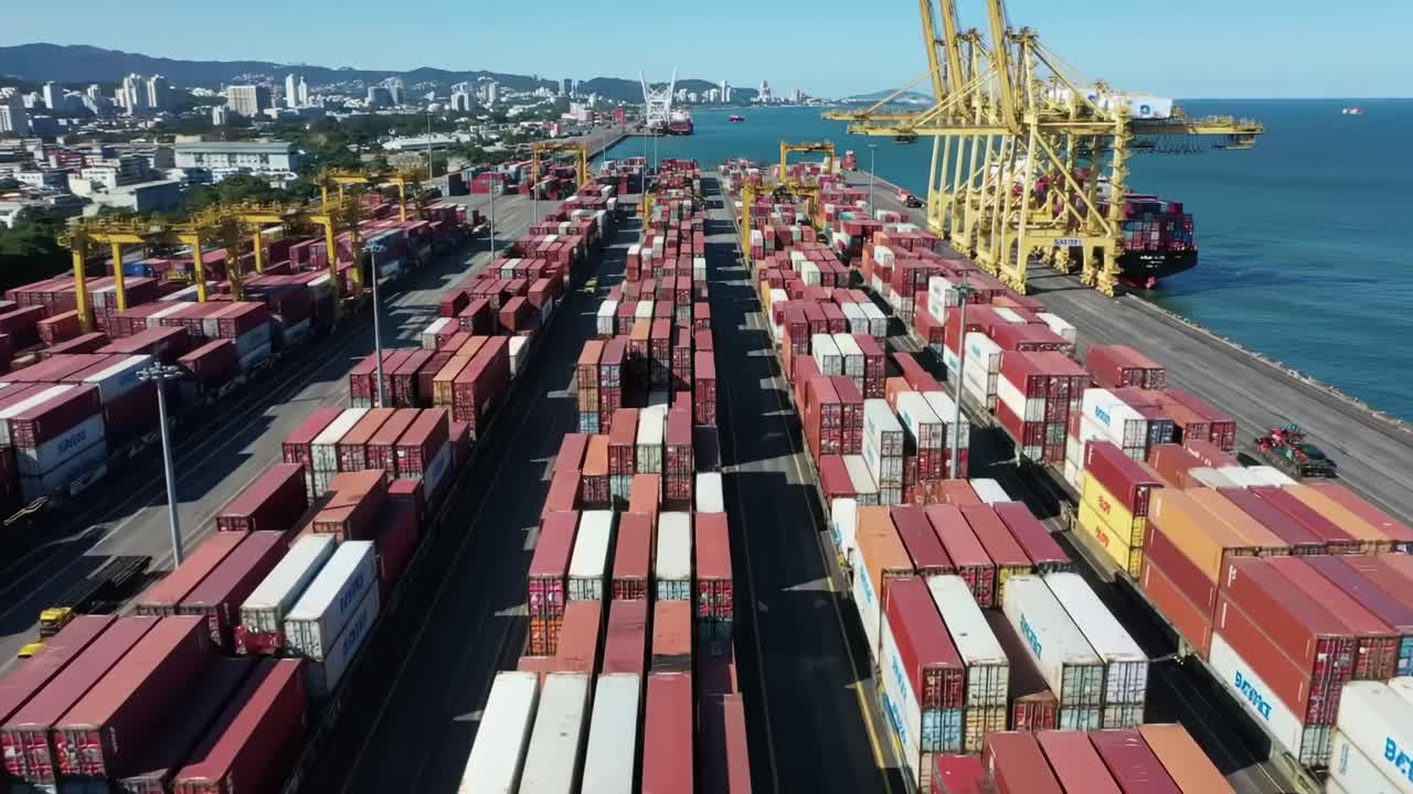 Containers are stacked at a bustling port with cranes loading ships in the background. The coastal city skyline provides a vibrant backdrop to this busy shipping hub.