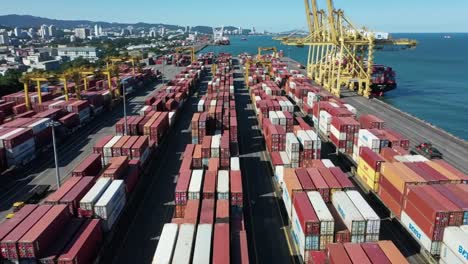 containers are stacked at a bustling port with cranes loading ships in the background. the coastal city skyline provides a vibrant backdrop to this busy shipping hub.