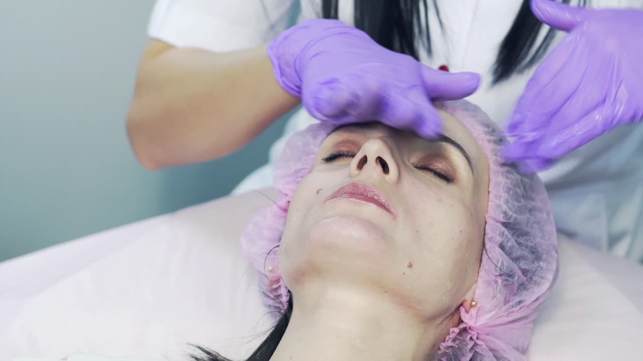 Beautiful woman is getting skin treatment and massage on her face with female hands in gloves at beauty cabinet