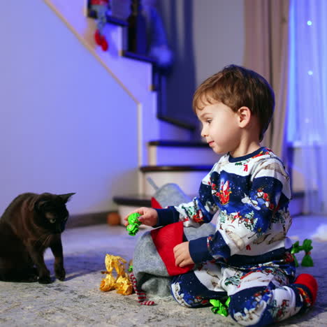small caucasian kid sits on the floor near christmas tree. toddler pulls the candies from stocking and puts them in front of the black cat.